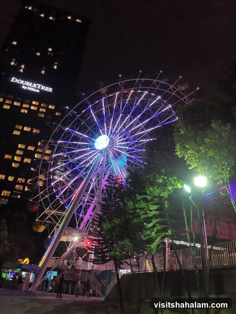 A brightly lit Ferris wheel is the central focus of this nighttime scene. The wheel is adorned with colorful LED lights in shades of purple, blue, pink, and white, creating a vibrant spectacle against the dark sky. To the left, a tall building with illuminated windows is visible, bearing the "DOUBLETREE by Hilton" logo. In the foreground and to the right, trees and park-like elements are present, with some people visible near the base of the Ferris wheel, suggesting an amusement park or recreational area.