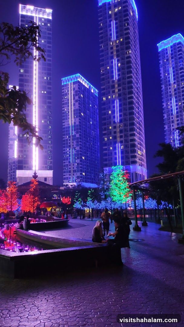 A nighttime scene featuring several tall, modern buildings illuminated with vibrant blue and white lights. In the foreground, a park area is decorated with trees covered in colorful lights, including red, green, and blue. A water feature with purple lights is visible, and several people are seated or walking in the park. The ground is paved with textured stones.