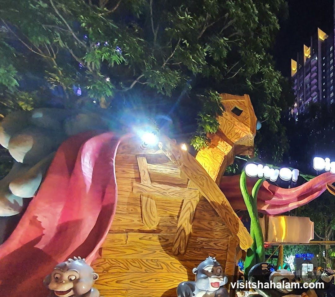 A nighttime view of a wooden treehouse-themed attraction with a red slide and cartoon animal figures. The attraction is illuminated by spotlights and string lights. In the background, a modern high-rise building is visible.