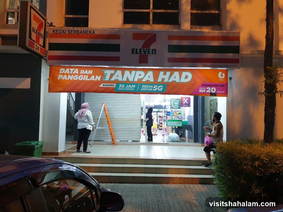 The exterior of a 7-Eleven convenience store at night. A large sign above the entrance reads "ELEVEN" with horizontal colored stripes. Below this, an orange banner advertises "DATA DAN PANGGILAN TANPA HAD" (Data and Calls Unlimited) with details about 30GB data and 5G. Another promotion for "PRABAYAR U25" offers a plan for RM20/bulan. Two individuals are visible near the entrance, one standing on a ladder and another inside the store. A car is parked in front of the store.