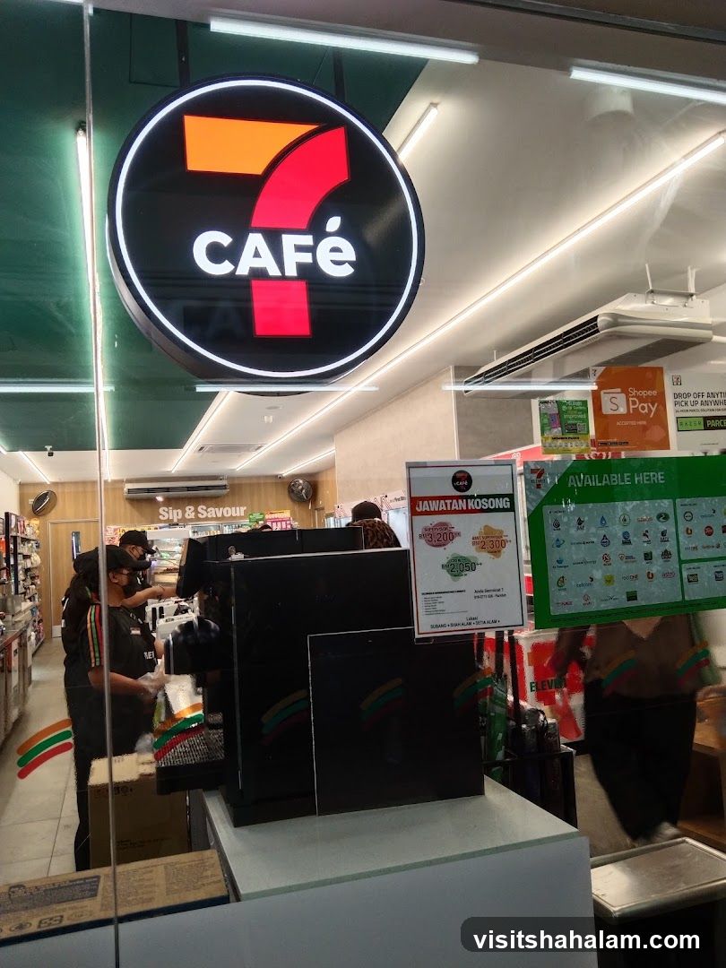 The image shows the entrance to a 7-Eleven store with a prominent "7 CAFÉ" sign. Inside, staff members are visible behind a counter, and various promotional materials and payment options like ShopeePay are displayed. The store appears to be a convenience store offering food and beverages.