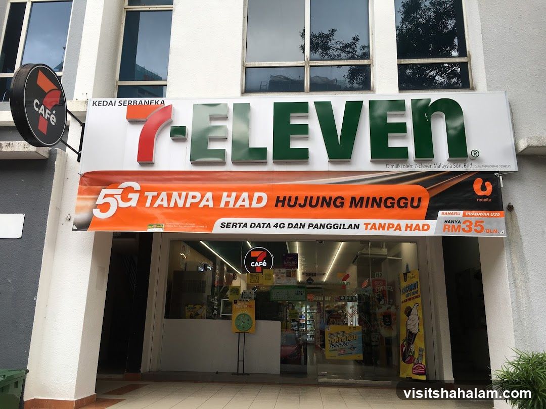The exterior of a 7-Eleven convenience store. A large sign with the 7-Eleven logo is prominently displayed above the entrance. Below the main sign, an orange banner advertises a "5G TANPA HAD HUJUNG MINGGU" promotion with U Mobile. The store entrance features glass doors and windows, revealing shelves stocked with products inside. A circular "CAFÉ" sign is visible to the left of the main 7-Eleven sign.