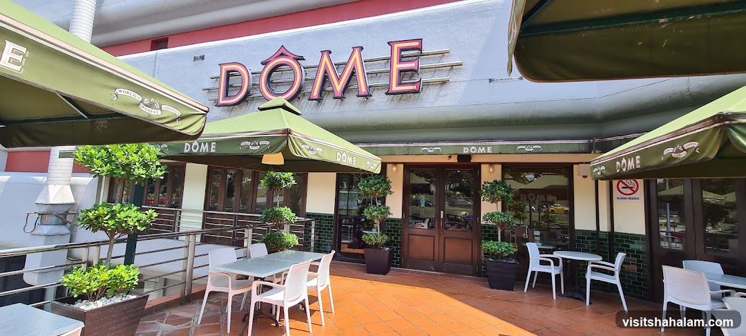 The exterior of a Dome cafe with outdoor seating. The cafe has a large sign with the word "DOME" in prominent lettering above the entrance. Green awnings with the cafe's name and "World's Finest Coffee" are visible. The entrance features double wooden doors and large windows. Outdoor tables and chairs are set up on a tiled patio, surrounded by potted plants.