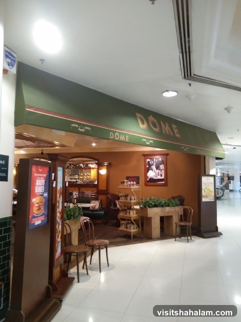 The exterior of a Dome cafe with a green awning that has the cafe's name, DOME, written in gold lettering. The entrance is visible, with seating and displays inside. A promotional poster for a Wagyu Burger is displayed to the left of the entrance.
