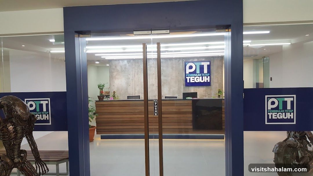 A view through glass double doors into an office reception area. The doors have a dark blue frame and wooden handles. A sign with the PTT logo and text "PEMBINAAN TETAP TEGUH" is visible on the wall behind the reception desk. The reception desk is made of wood slats and has a dark counter. There are plants on the desk and in pots. To the left and right of the entrance, on the glass, are similar PTT logos. A metal sculpture of a figure is visible to the left, and a sculpture of a motorcycle is visible to the right.