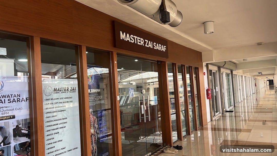 The exterior of a business with a wooden frame and glass doors. A sign above the entrance reads "MASTER ZAI SARAF". A poster on the left window lists services such as "SAKIT BELAKANG (OTOT MENGERAS)", "SLIP DISC (HNP ATAU BULDING DISC)", and "UPPER/LOWER BACKPAIN".