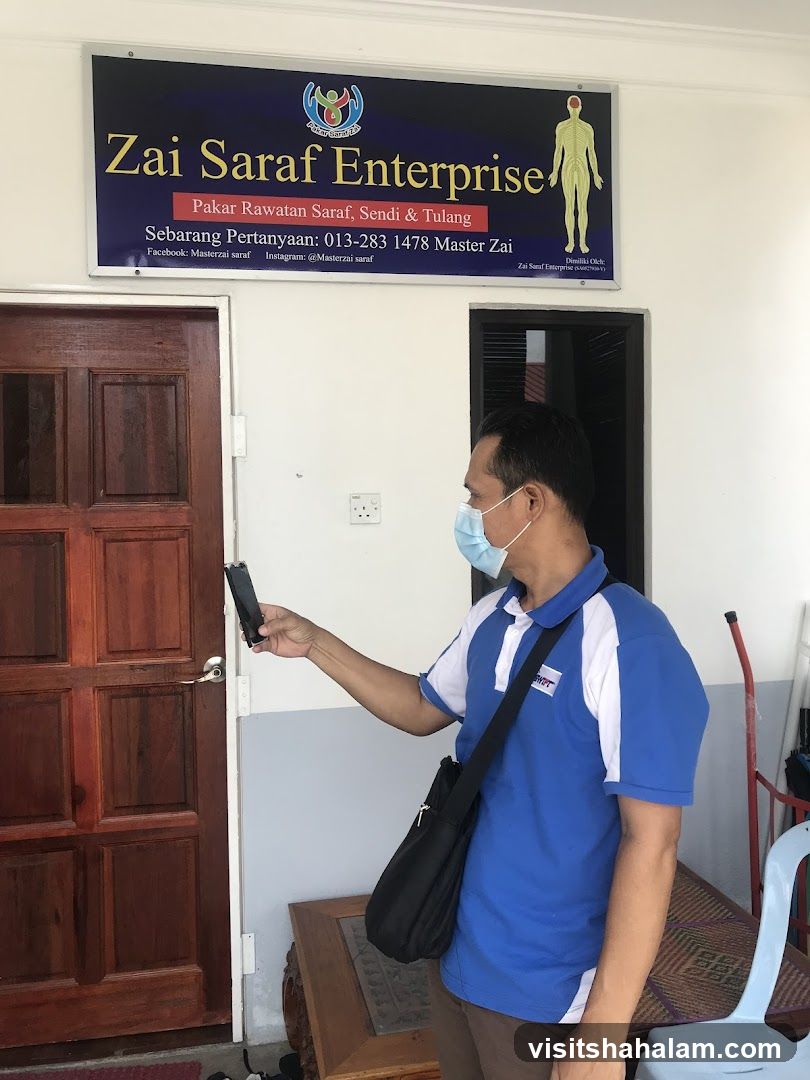 A man wearing a blue and white polo shirt and a face mask is standing in front of a wooden door. Above the door is a sign that reads "Zai Saraf Enterprise" with text below detailing services and contact information. The sign also features a logo and an illustration of the human nervous system.