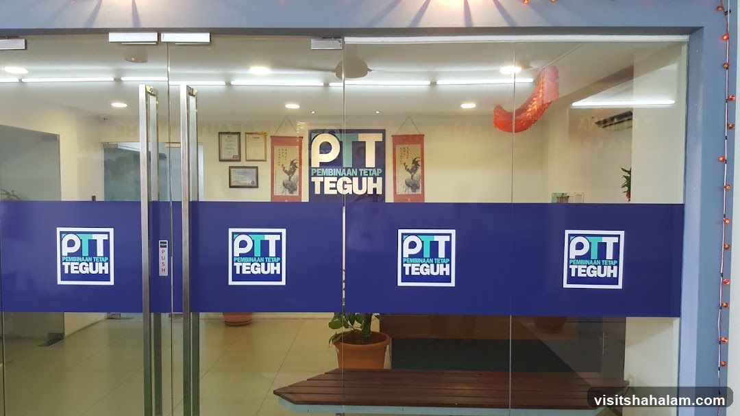The image shows the glass entrance of a business with multiple logos and signage visible. The main signage on the glass doors and windows reads "PTT PEMBINAAN TETAP TEGUH" in a blue and white square logo. Above the doors, a larger version of the same logo is displayed. Inside, framed certificates and decorative scrolls are visible on the wall. A red dragon decoration hangs from the ceiling. A wooden bench and potted plants are also present.