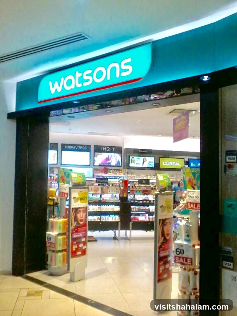 The entrance to a Watsons store is shown. A large, illuminated sign above the entrance reads "watsons". Inside the store, shelves are stocked with various beauty and health products. Promotional displays and sale signs are visible near the entrance.