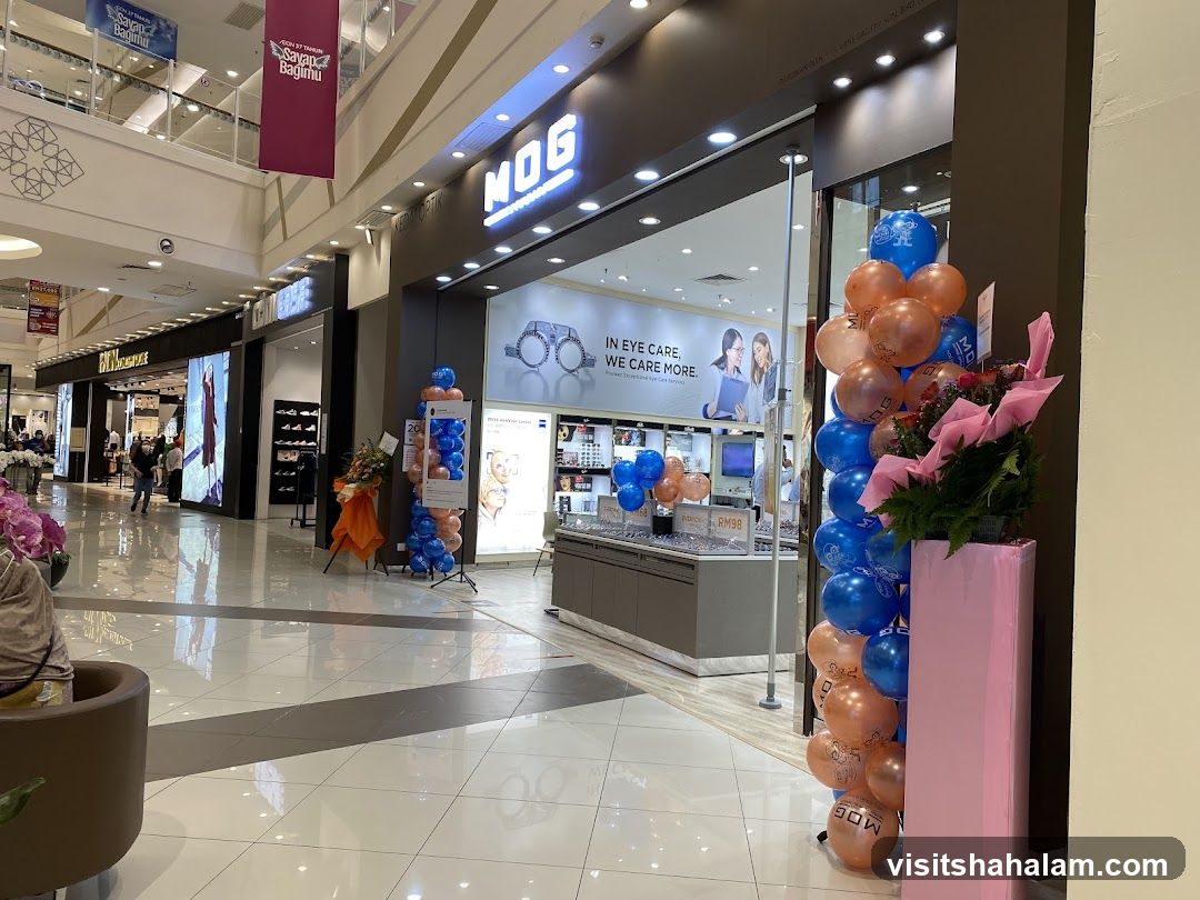 The storefront of an optometrist store named MOG. The store features a large illuminated sign with the MOG logo. Inside, displays of eyeglasses and eye care services are visible. A large balloon arrangement and floral display are placed outside the entrance. The store is located within a shopping mall, with other shops visible in the background.
