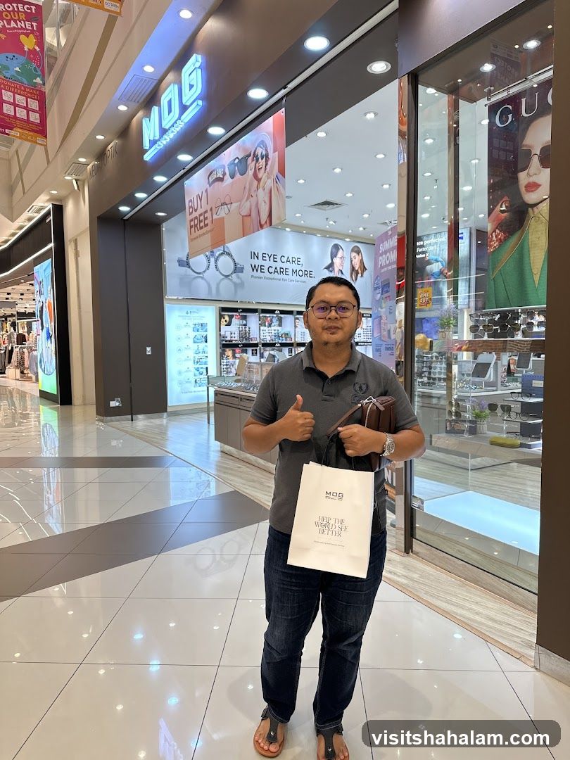 A man stands in front of an MOG Optics store, giving a thumbs up. The store has a large sign that says "MOG OPTICS" and features advertisements for "BUY 1 FREE 1" and "IN EYE CARE, WE CARE MORE." The store interior displays various eyewear. A shopping bag with the MOG logo is visible.