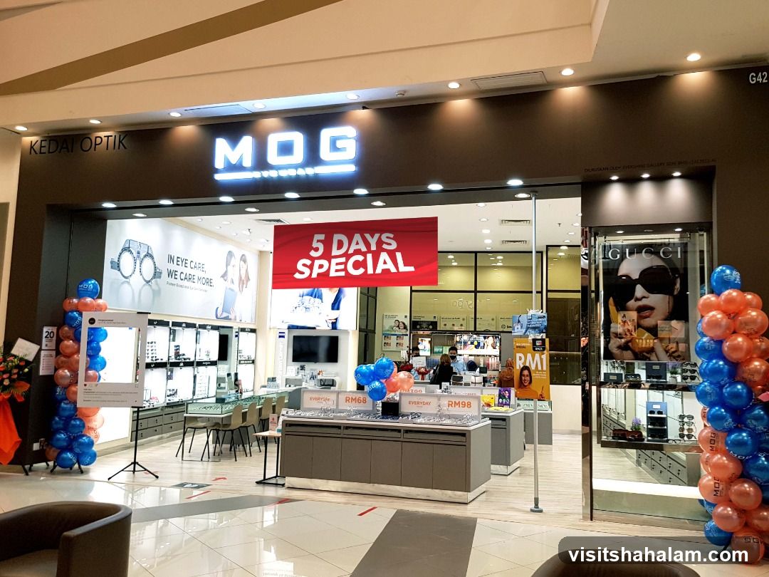 The exterior of an MOG Eyewear store, also labeled as KEDAI OPTIK. The store features a large illuminated sign that reads "MOG EYEWEAR". Inside, displays of eyeglasses and sunglasses are visible. A prominent red banner announces a "5 DAYS SPECIAL". To the right, a large advertisement for Gucci sunglasses is displayed. The entrance is decorated with balloons in blue and orange.