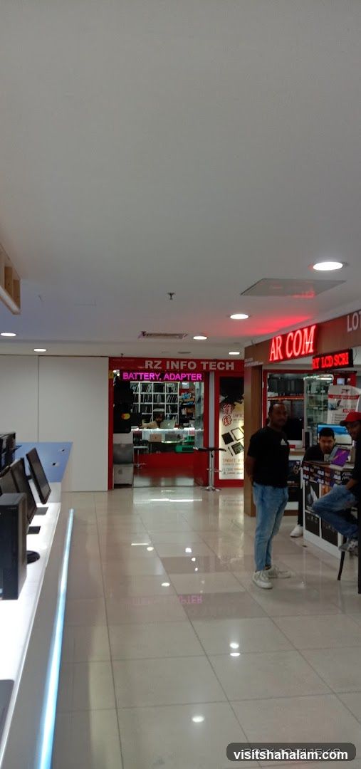 The storefront of RZ INFO TECH, a shop selling batteries and adapters. The shop has a red facade with a sign above the entrance. Inside, shelves are stocked with various electronic components. To the right, another shop, AR COM, is visible, displaying electronic items.