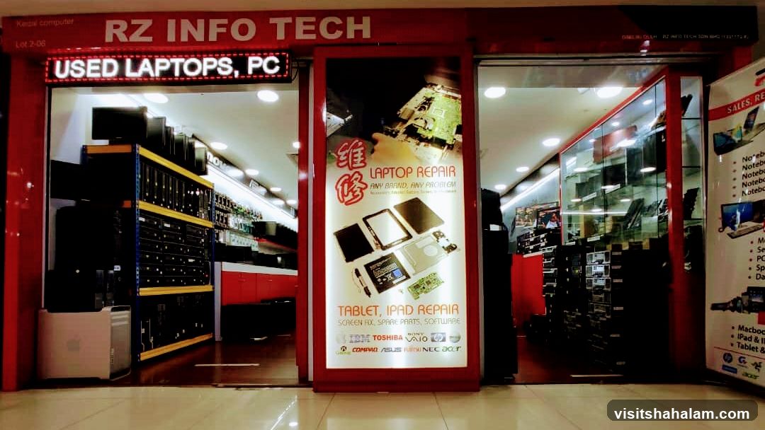 The storefront of RZ INFO TECH, a computer repair and sales shop. The shop has a red facade with the business name prominently displayed. Inside, shelves are filled with computers and laptops. A large illuminated sign advertises laptop and tablet repair services, listing brands like IBM, Toshiba, Sony, Compaq, Asus, and NEC. Another sign indicates they sell used laptops and PCs.