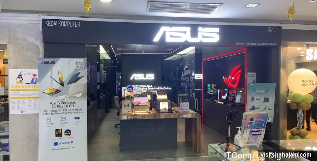 The exterior of an ASUS computer store, labeled 'KEDAI KOMPUTER'. The store features a large illuminated ASUS logo above the entrance. Inside, various ASUS laptops and computer components are displayed. A promotional banner for the ASUS Zenbook 14 Flip OLED is visible outside the store, along with a sign for 'C-ZONE JOIN OUR TEA'. A large balloon with 'Galaxy A34 A54 5G' is also present.