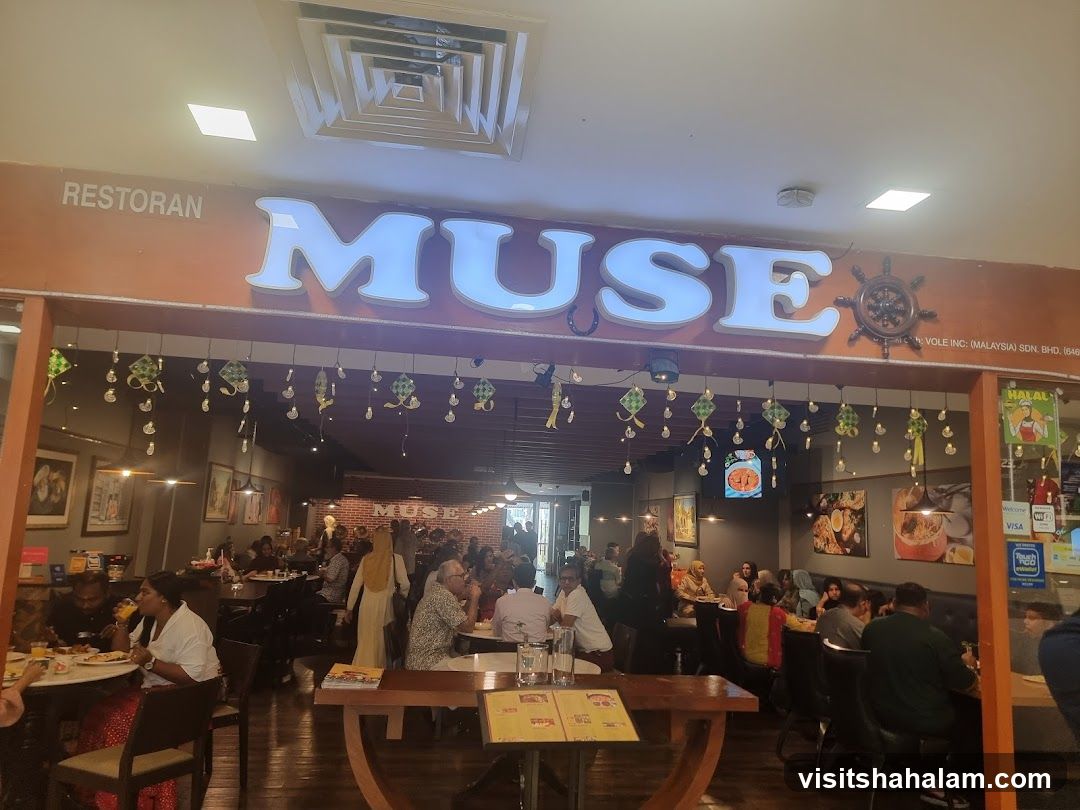 The exterior of a restaurant called Muse, with large illuminated letters spelling out the name. The restaurant appears to be busy with patrons dining inside. Decorative elements, possibly for a festival, hang from the ceiling.