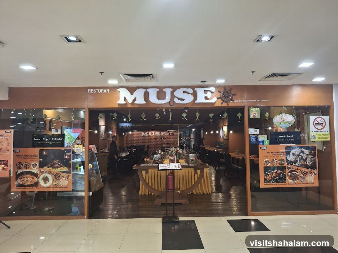 The exterior of a restaurant called Muse, with large illuminated letters spelling out the name above the entrance. The restaurant has a warm, inviting interior visible through the glass facade, featuring wooden tables and chairs, and brick walls. Promotional posters showcasing food are displayed on the left and right sides of the entrance.