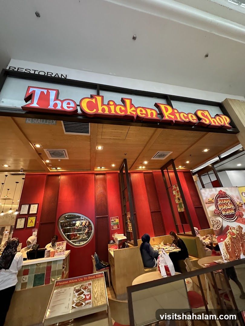 The exterior of a restaurant called "The Chicken Rice Shop". The sign is large and red with yellow trim. Inside, the restaurant has red walls and wooden booths. Several customers are seated at tables.
