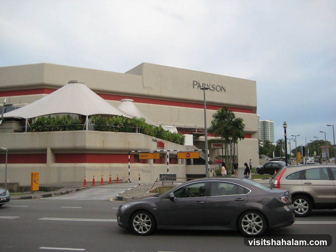 A wide shot of the exterior of a Parkson department store. The building is large and modern, with a beige facade and red trim. The word "PARKSON" is prominently displayed in large letters on the upper part of the building. In front of the building, there are several cars on the road and a few pedestrians walking. The sky is overcast.
