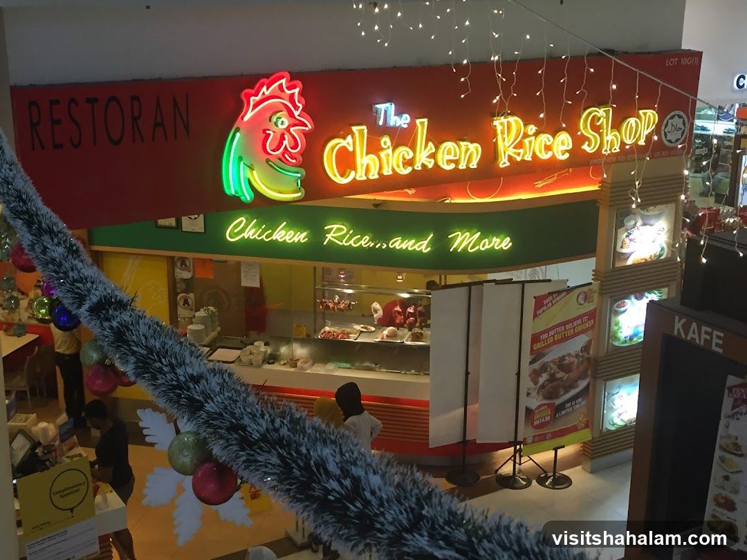 A neon sign for "The Chicken Rice Shop" is visible above the entrance of a restaurant. The sign features a cartoon chicken and the words "Restoran" and "Chicken Rice...and More". There are also promotional posters and displays inside and outside the shop.