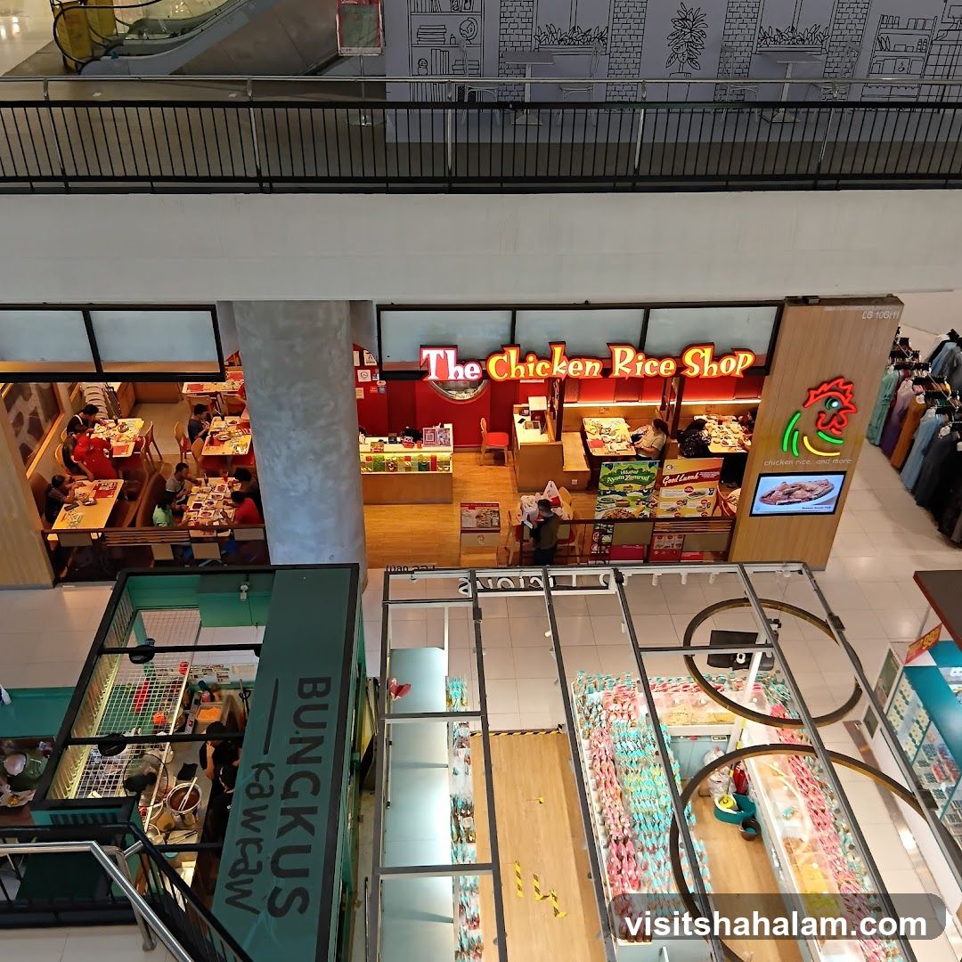 An aerial view of a brightly lit restaurant called "The Chicken Rice Shop" inside a mall. The shop has a red and wooden facade with a prominent neon sign. Inside, customers are seated at tables, and staff are visible behind the counter. Various food displays and promotional materials are visible within the shop.