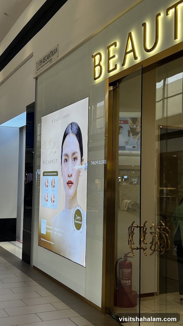 The exterior of a beauty salon with a large illuminated sign that reads "BEAUTÉ" in gold lettering. To the left, a large digital display shows a woman's face and text related to a treatment called "PicoSpot" with "Instant Glow-Up in 1 Session" and "Zero Downtime". The entrance has ornate gold handles and a fire extinguisher is visible inside.