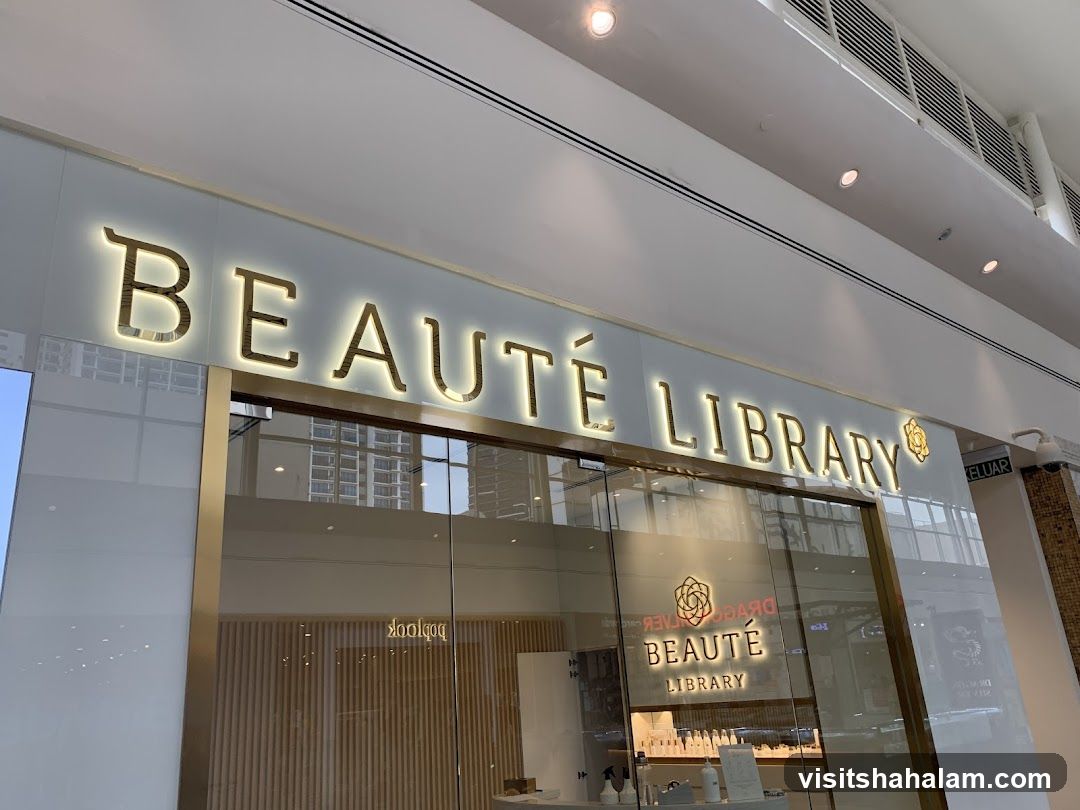 The exterior of a Beauté Library shop. The shop has a large, illuminated sign with the name "BEAUTÉ LIBRARY" in gold lettering. Below the sign, there are large glass windows and doors, revealing the interior of the shop. The interior appears to be modern and clean, with shelves displaying various beauty products.