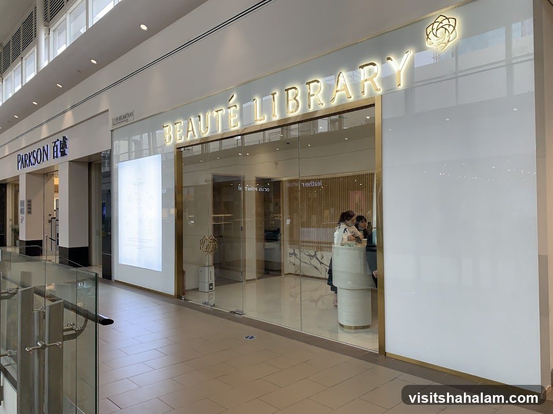The exterior of a Beauté Library shop with large glass windows and doors. The shop's name is displayed in gold lettering above the entrance. Inside, two staff members are visible behind a counter. To the left of the shop, a Parkson store is visible.