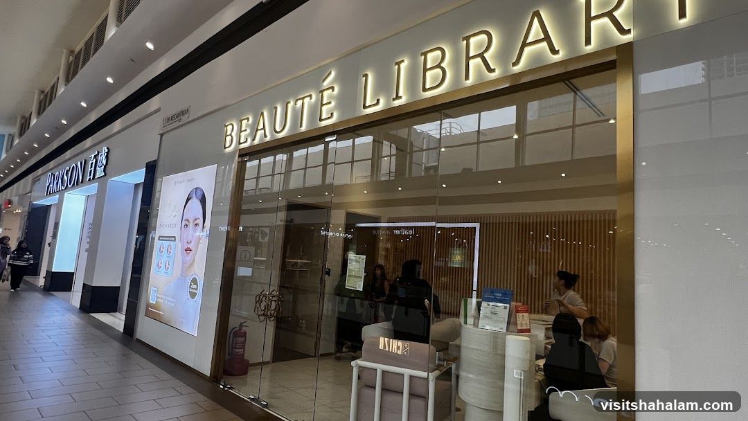 The exterior of a beauty store called Beauté Library. The store has a large glass storefront with the name illuminated in gold lettering above the entrance. Inside, there are several people and displays visible. To the left of Beauté Library is another store with the name Parkson.
