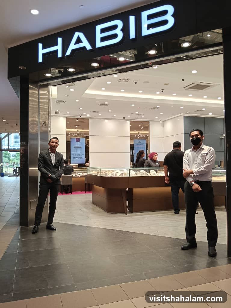 The exterior of a Habib jewelry store with the store name prominently displayed in large white letters on a black background above the entrance. Two men in suits stand at the entrance, one on the left and one on the right. Inside, display cases filled with jewelry are visible, and other staff members are attending to customers.