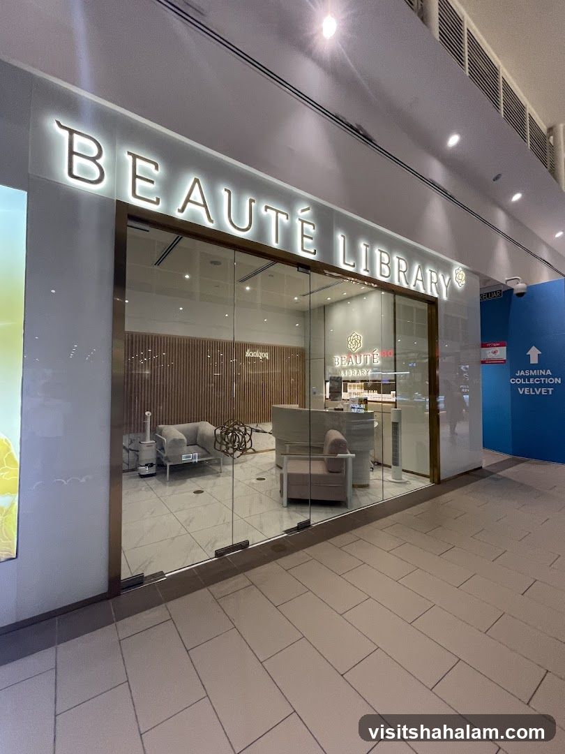 The exterior of a modern beauty store with a large glass facade. The store's name, "BEAUTÉ LIBRARY", is prominently displayed in illuminated lettering above the entrance. Inside, the store appears to be well-lit and features minimalist decor, including seating areas and display shelves.