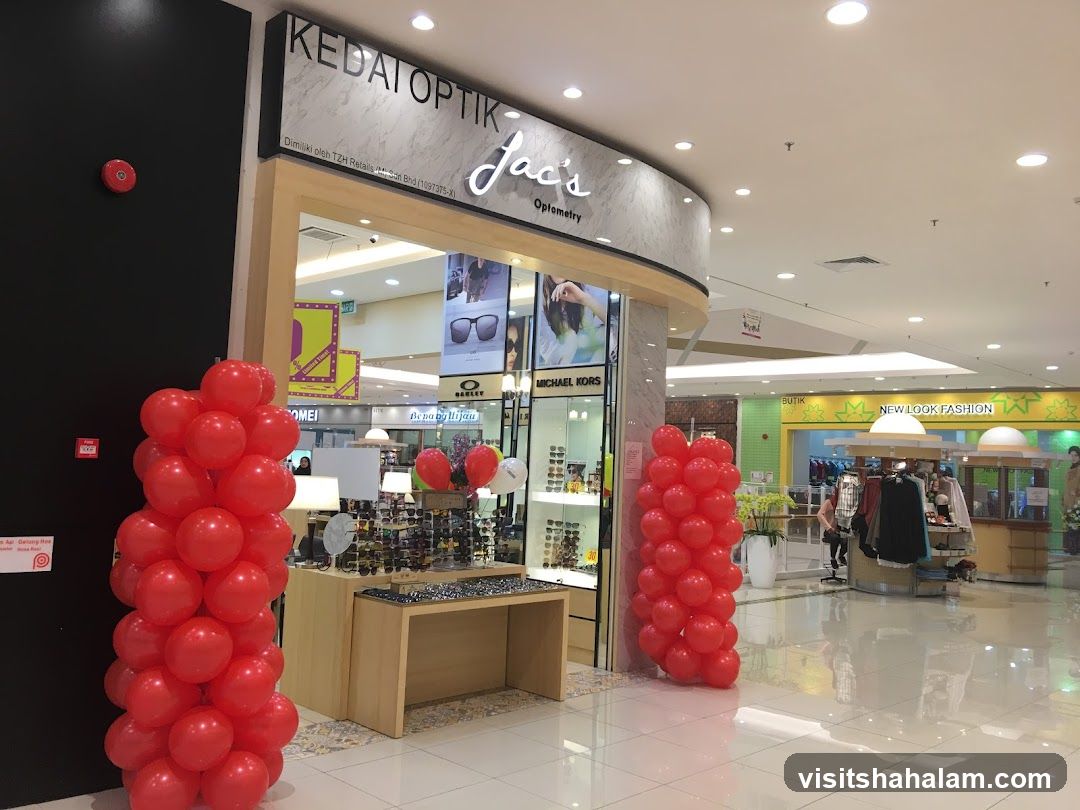 The exterior of a shop called Jac's Optometry, which sells eyewear. The shop front is decorated with red balloons. Inside, displays of glasses and sunglasses are visible. Other shops and a mall interior are visible in the background.