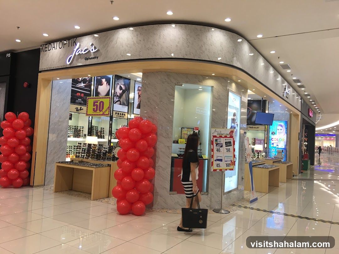 The exterior of an optometry shop with a prominent sign that reads "KEDAI OPTIK Jac's Optometry". The storefront displays various eyewear and promotional materials. Red balloons are arranged on either side of the entrance.