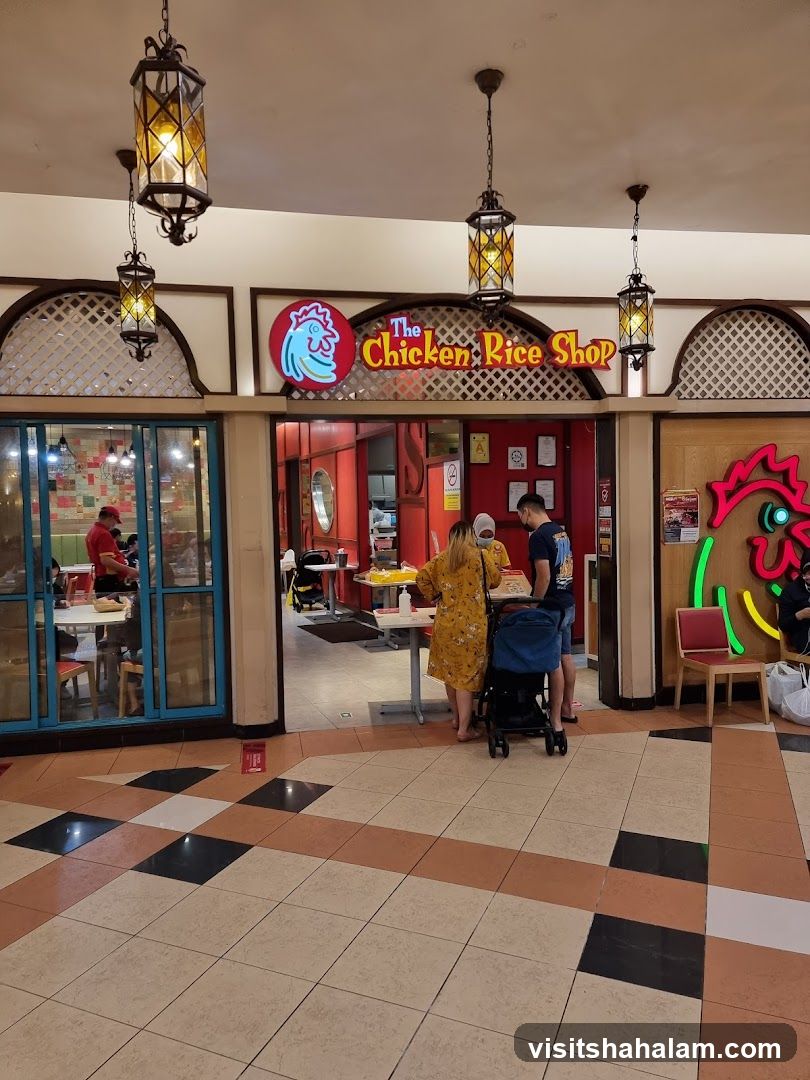 The exterior of a restaurant called "The Chicken Rice Shop". The entrance is open, and people are seen inside and at the counter. There are tables and chairs visible inside. The shop has a red and white color scheme with a prominent chicken logo.