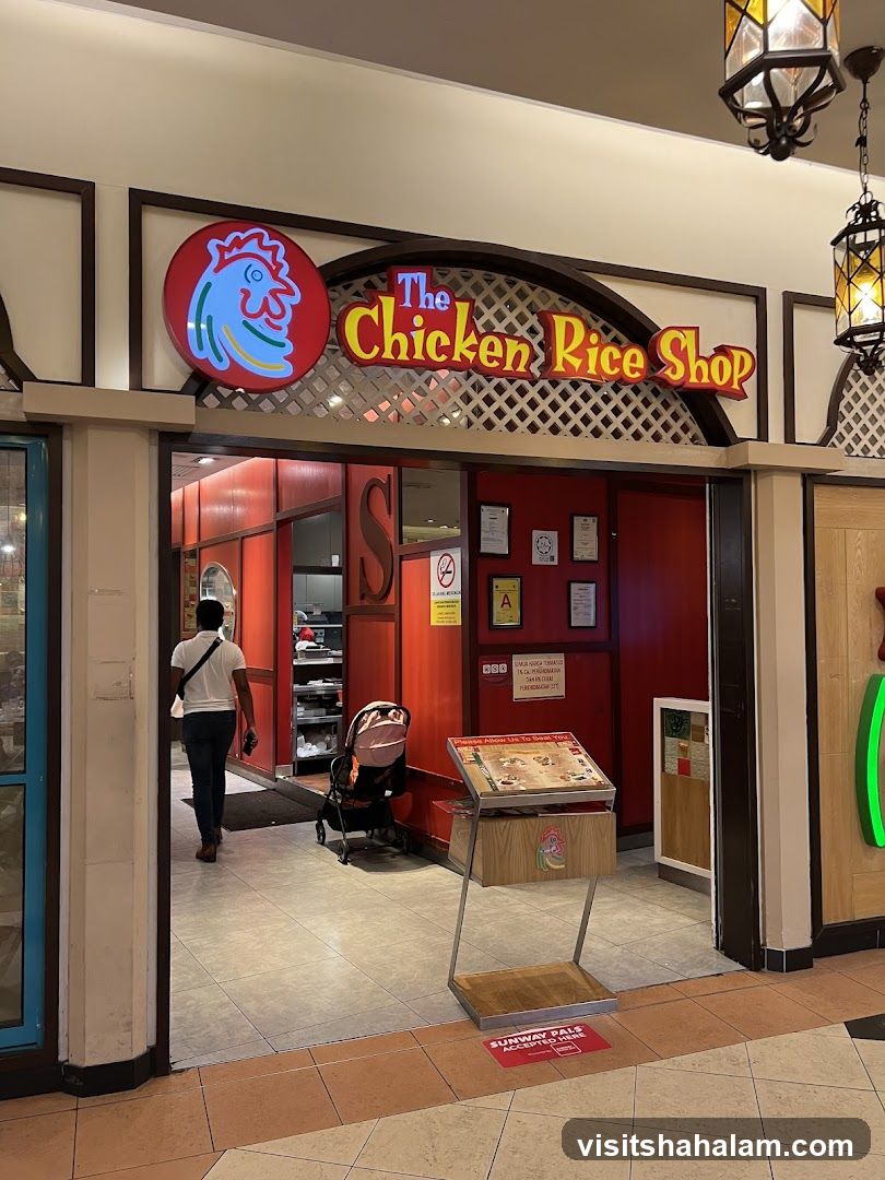 The exterior of a restaurant called "The Chicken Rice Shop". The entrance is wide and open, with red walls and decorative lattice work above the doorway. A sign with a cartoon chicken head and the restaurant name is prominently displayed. A man is walking into the restaurant, and a baby stroller is parked nearby. A menu stand is placed outside the entrance.