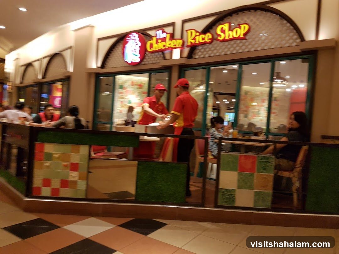 The exterior of a restaurant called "The Chicken Rice Shop" with a red and yellow sign above the entrance. People are seated inside and outside the restaurant.