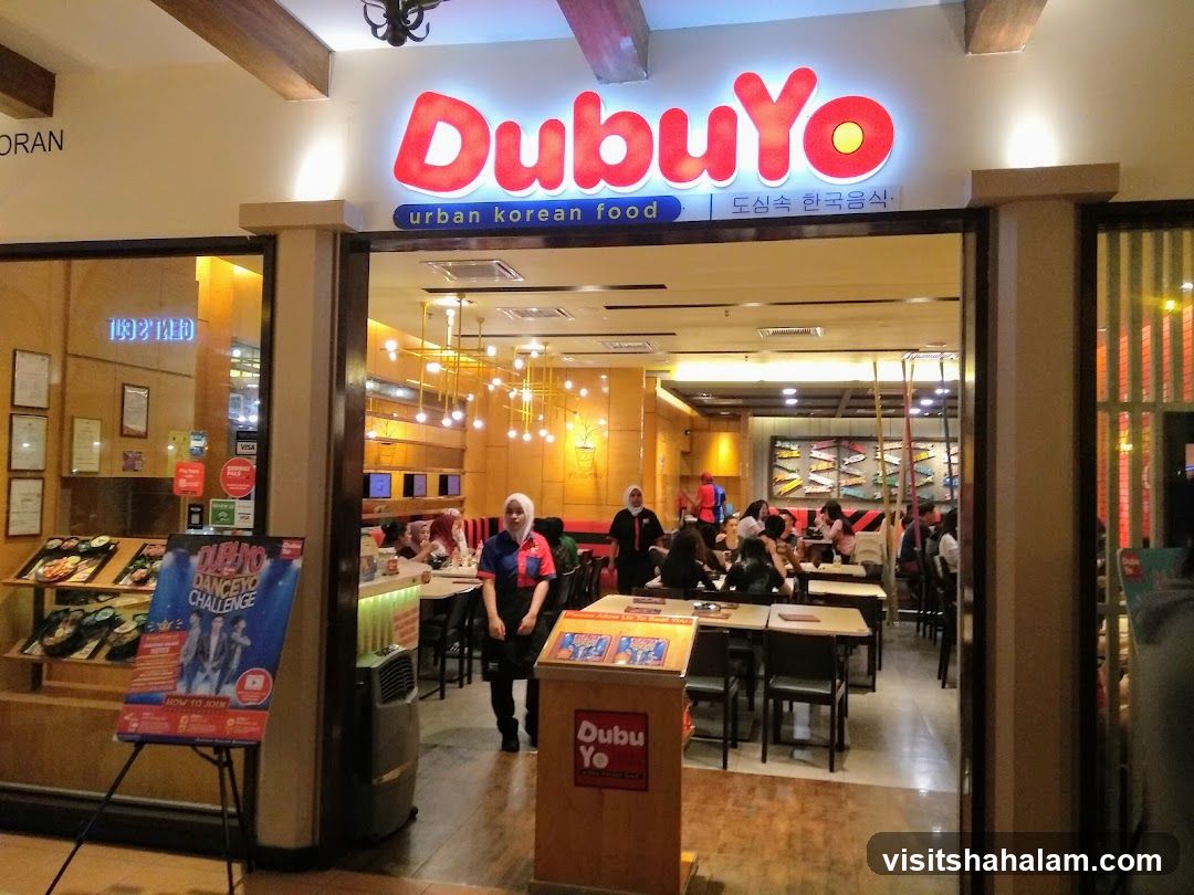 The exterior of a DubuYo restaurant, an urban Korean food establishment. The entrance is open, revealing a dining area with tables and chairs. Several patrons are seated and dining. A promotional poster for a "DubuYo Danceyo Challenge" is displayed outside the entrance.