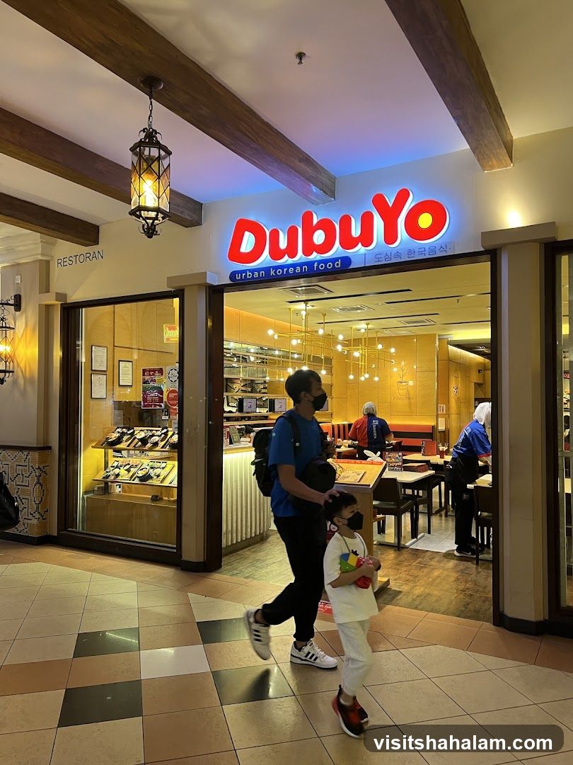 The exterior of a DubuYo restaurant, a Korean food establishment. The prominent red and yellow sign with the name "DubuYo" is visible above the entrance. Below the name, it reads "urban korean food" in English and Korean script. The restaurant interior, with tables and chairs, is visible through the glass facade. People are seen walking past the entrance and inside the restaurant.